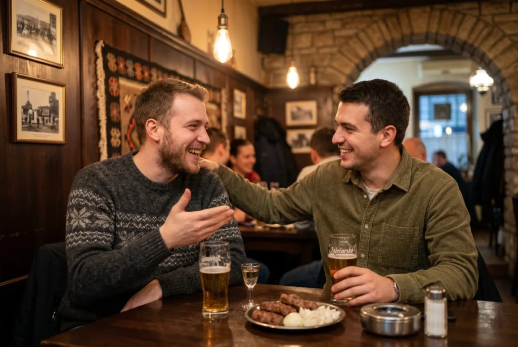 Dva prijatelja sede za drvenim stolom u kafani, razgovaraju uz pivo, rakiju i tanjir kobasica sa lukom.